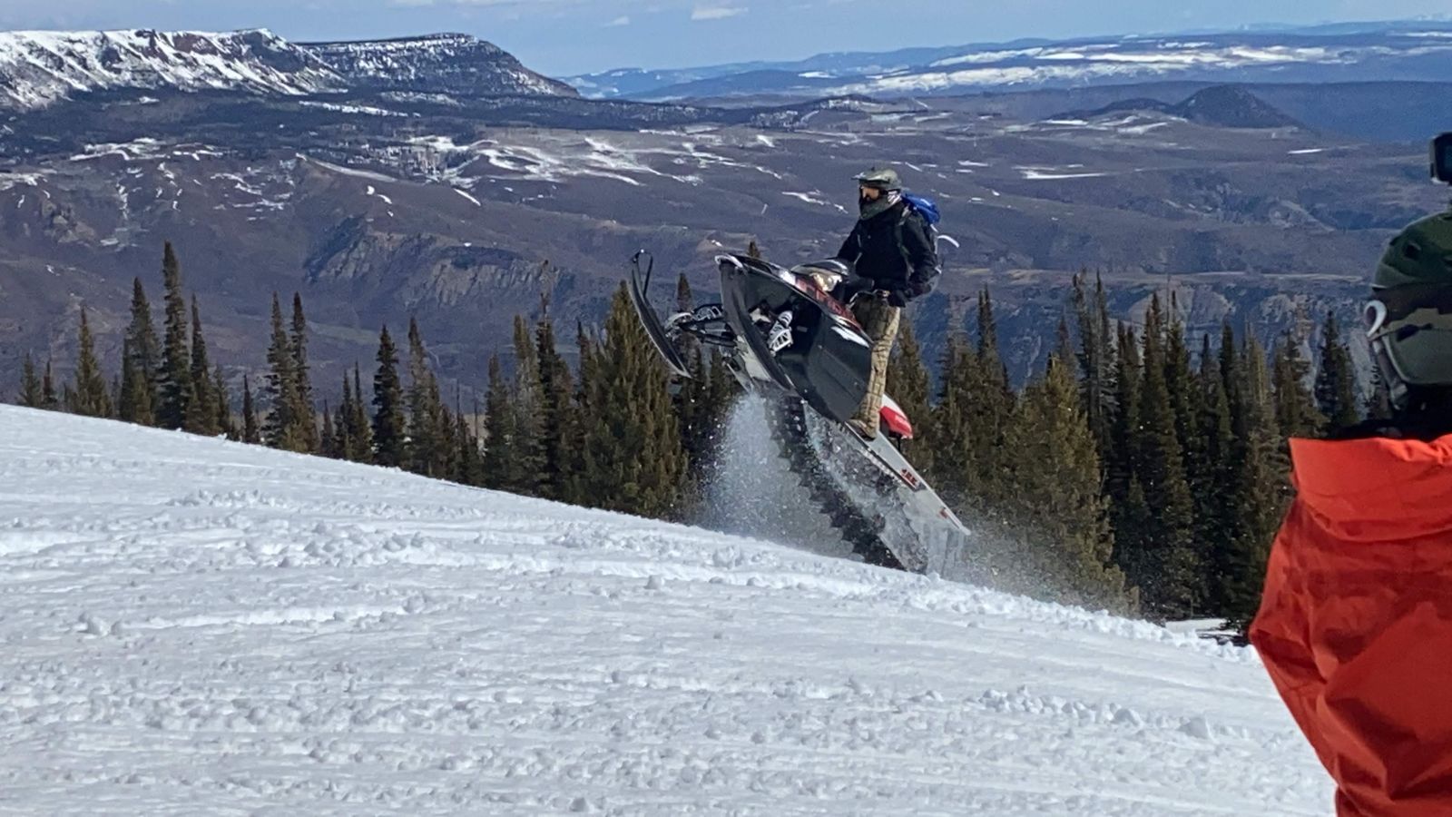 Rider on a dark snowmobile mid-jump, kicking up snow above evergreens and the wide valleys of the Flat Tops