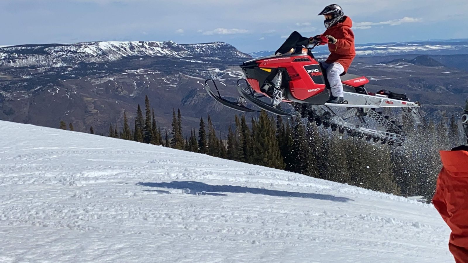 Rider in a red jacket catching air on a red snowmobile above snowy ridges and evergreens in Colorado's Flat Tops